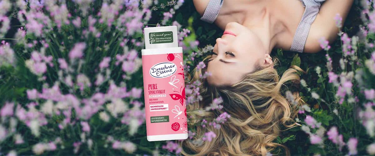 Frau liegt seitlich im Gras zwischen lila Blüten, neben ihr steht eine rosa Flasche Dresden Essence „Pure Sinnlichkeit“ Schaumbad mit sichtbarem Markenlogo und Aufschrift
