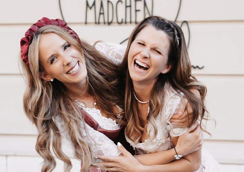 Zwei junge Frauen in hellen Festkleidern lachen und umarmen sich, hinter ihnen teilweise lesbar das Wort „Mädchen“ auf einer Wand