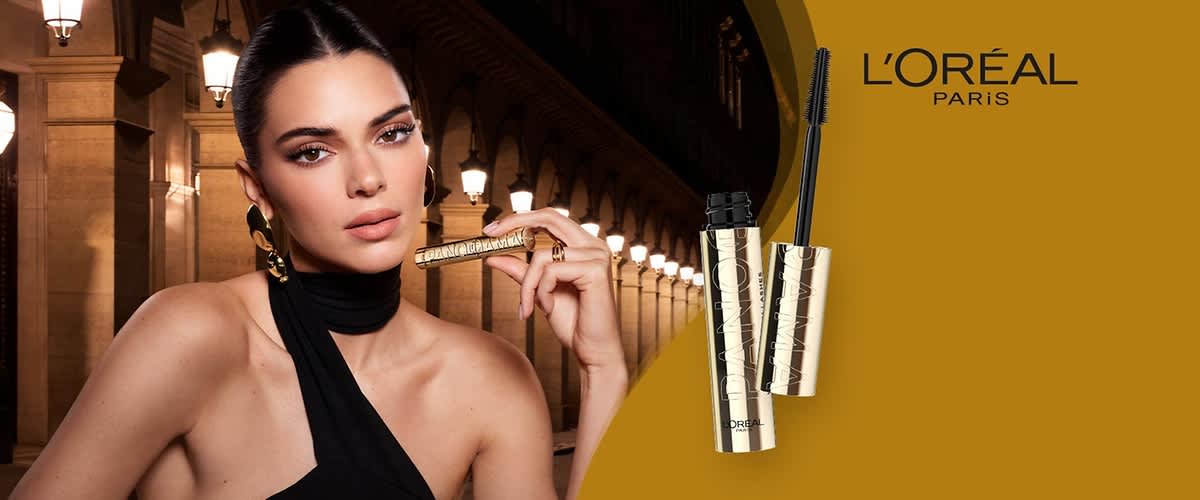 Eine goldene Mascara-Flasche mit schwarzem Bürstchen, darauf die Aufschrift „L'Oreal“. Im Hintergrund das Logo von L'Oréal Paris.