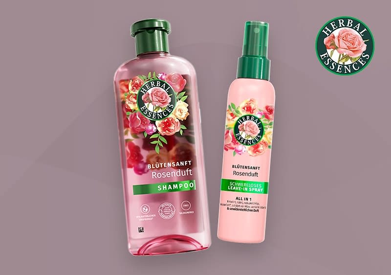 Zwei Haarpflegeprodukte der Marke Herbal Essences mit Rosenduft, links eine große durchsichtige Flasche mit rosa Flüssigkeit und der Aufschrift Blütensanft Rosenduft Shampoo sowie rechts ein kleineres rosa Spray mit der Aufschrift Blütensanft Rosenduft Glanzpflegespray, beide Produkte tragen das Herbal Essences Logo mit einer Rose und grünen Blättern, im Hintergrund ein grauer Farbverlauf und oben rechts das Herbal Essences Logo mit einer Rose und der Schrift Herbal Essences in einem grünen Kreis.