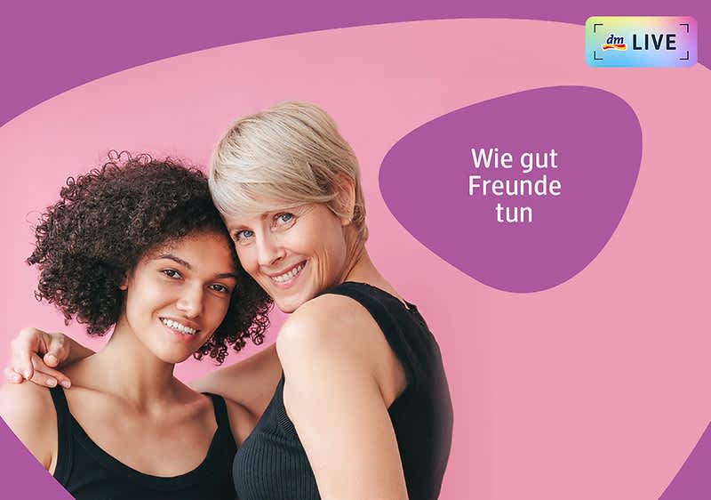 Zwei Frauen stehen nebeneinander vor einem rosa Hintergrund, die linke Frau hat lockiges dunkles Haar und trägt ein schwarzes ärmelloses Oberteil, die rechte Frau hat kurzes blondes Haar und trägt ebenfalls ein schwarzes ärmelloses Oberteil, beide haben ihre Gesichter verschwommen, oben rechts steht in einem lila Bereich der Text Wie gut Freunde tun und daneben ist das dm LIVE Logo mit einem kleinen Regenbogenrahmen sichtbar