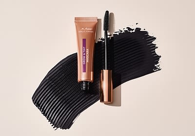 Eine Tube mit der Aufschrift „Dr. Hauschka Magic Finish“ und „CASHMERE MATTE FOUNDATION“ in einem bronzefarbenen Design liegt neben einer geöffneten Mascara mit schwarzer Bürste, beide auf einem Hintergrund mit einem breiten Pinselstrich aus schwarzer Farbe.