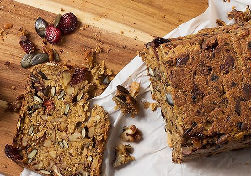 Ein angeschnittenes Vollkornbrot mit Nüssen und Samen liegt auf einem Holzbrett, daneben einige herausgefallene Zutaten wie Kürbiskerne und getrocknete Früchte