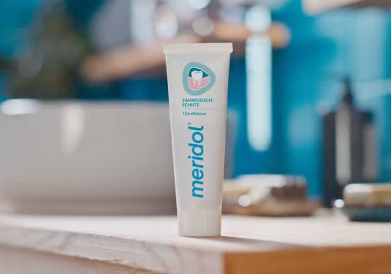 Eine weiße Tube Meridol Zahnpasta mit blauem Logo und kleinem runden Siegel vorne steht aufrecht auf einem hellen Badewaschtisch im Badezimmer