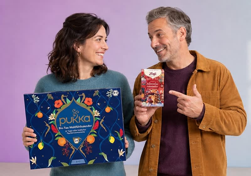 Frau und Mann lächelnd, die Frau hält einen blauen Pukka Tee-Wohlfühlkalender, der Mann zeigt auf eine rote Yogi Teebox mit der Aufschrift „Ayurvedic Amaretto“. Bild mithilfe von KI erstellt
