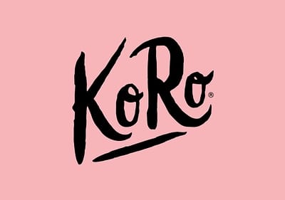 Das Bild zeigt das Logo der Marke KoRo in schwarzer Schrift auf einem rosa Hintergrund, wobei das K und R in geschwungenen Großbuchstaben geschrieben sind und das o in Kleinbuchstaben, mit einem kleinen ®-Symbol rechts neben dem o