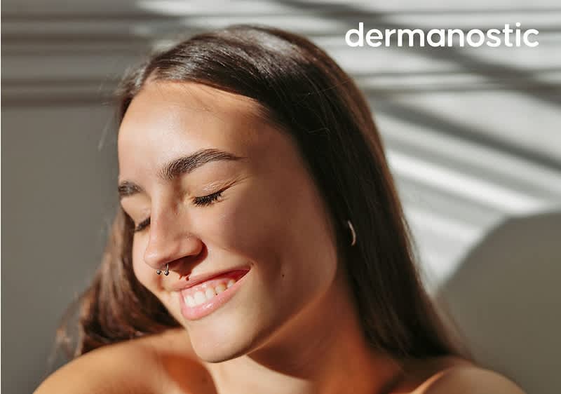 Junge Person mit geschlossenen Augen und leichtem Lächeln, sichtbarer Nasenpiercing und langes dunkles Haar, im Bild oben rechts Schriftzug „dermanostic“