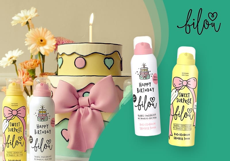Zwei Duschschaum-Flaschen der Marke Bilou mit den Aufschriften „Happy Birthday“ und „Sweet Surprise“ vor einem grünen Hintergrund, daneben eine mehrstöckige Torte mit rosa Schleife und Kerze
