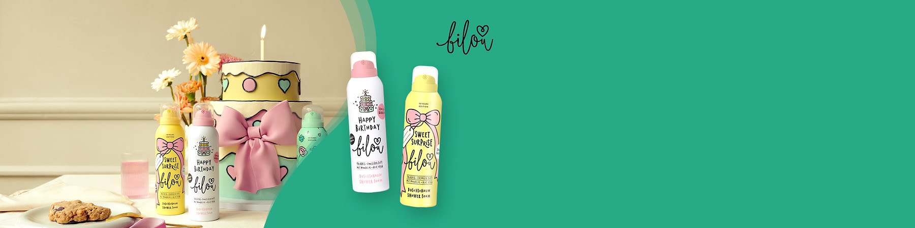 Zwei Duschschaumflaschen der Marke Bilou, eine in Rosa mit der Aufschrift „Happy Birthday“ und eine in Gelb mit der Aufschrift „Sweet Surprise“, daneben eine gelbe Geburtstagstorte mit rosa Schleife und Blumen im Hintergrund