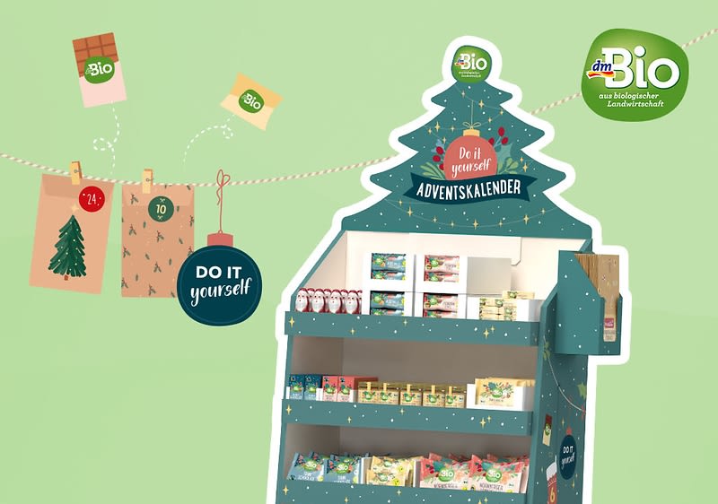 Ein grüner Adventskalender in Form eines Tannenbaums mit der Aufschrift Do it yourself Adventskalender und dem dmBio-Logo oben, darunter sind verschiedene Bio-Produkte wie Tees und Snacks sichtbar, daneben hängt eine Girlande mit einem roten Kreis mit der Aufschrift Do it yourself, einem grünen Quadrat und einem braunen Umschlag mit der Zahl 24, im Hintergrund ist ein hellgrüner Farbverlauf zu sehen