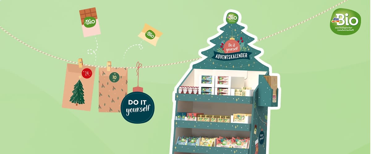 Ein grüner Adventskalender in Form eines Tannenbaums mit der Aufschrift Do it yourself Adventskalender und dem dmBio-Logo oben, darunter sind verschiedene Bio-Produkte wie Tees und Snacks sichtbar, daneben hängt eine Girlande mit einem roten Kreis mit der Aufschrift Do it yourself, einem grünen Quadrat und einem braunen Umschlag mit der Zahl 24, im Hintergrund ist ein hellgrüner Farbverlauf zu sehen