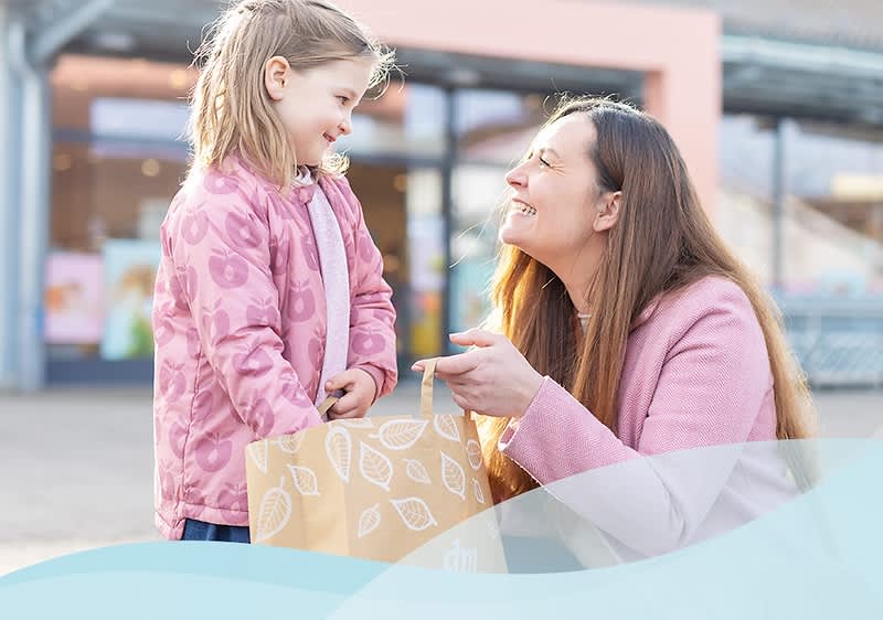 Eine Frau mit langen braunen Haaren und einem rosa Mantel kniet vor einem Kind, das ebenfalls einen rosa Mantel trägt, und hält eine Einkaufstasche mit Blattmuster in der Hand, während sie sich draußen vor einem dm-Markt befinden, dessen Eingang im Hintergrund zu sehen ist