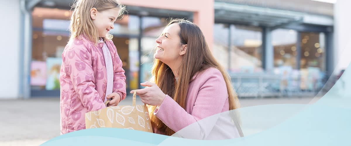 Eine Frau mit langen braunen Haaren und einem rosa Mantel kniet vor einem Kind, das ebenfalls einen rosa Mantel trägt, und hält eine Einkaufstasche mit Blattmuster in der Hand, während sie sich draußen vor einem dm-Markt befinden, dessen Eingang im Hintergrund zu sehen ist