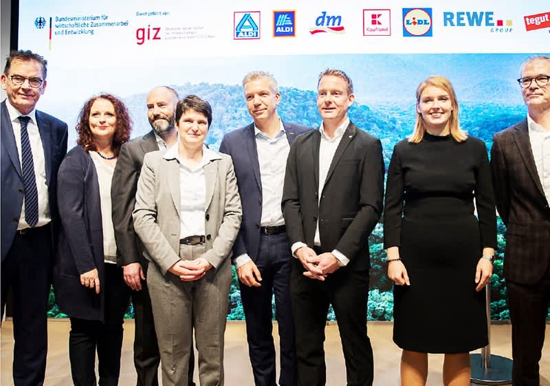 Eine Gruppe von acht Personen in formeller Kleidung steht nebeneinander vor einem Hintergrund mit einem Waldmotiv, über ihnen sind Logos verschiedener Unternehmen und Organisationen wie Aldi, dm, Lidl, Rewe und Tegut zu sehen, die zusammen mit dem Bundesministerium für wirtschaftliche Zusammenarbeit und Entwicklung und der Deutschen Gesellschaft für Internationale Zusammenarbeit (giz) auf einem Banner abgebildet sind.