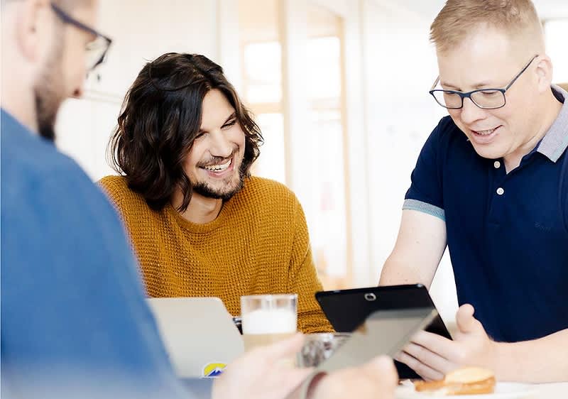 Drei Personen sitzen an einem Tisch und arbeiten gemeinsam, wobei zwei von ihnen Tablets in den Händen halten und eine Person vor einem Laptop sitzt, auf dem Tisch stehen ein Glas mit einem Getränk und ein Teller mit einem Gebäckstück