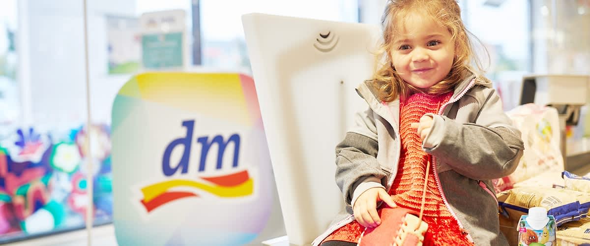 Ein kleines Mädchen mit langen Haaren sitzt auf einer Ladentheke und trägt ein rotes Kleid mit einer grauen Jacke darüber, hält eine kleine Tasche in der Hand und neben ihr steht eine Flasche mit einem Getränk, im Hintergrund ist ein Schild mit dem dm-Logo zu sehen