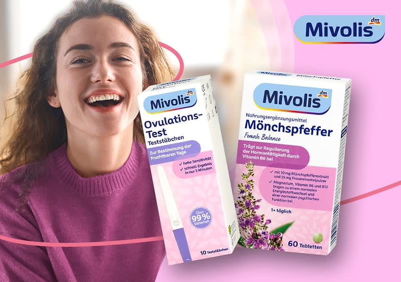 Frau in violettem Pullover hält links ein rosafarbenes Mivolis Ovulationstest-Teststäbchen-Packung, rechts daneben eine Mivolis Mönchspfeffer-Vitaminpackung mit lila Blumenmotiv und dem Markenlogo „Mivolis“ oben rechts im Bild