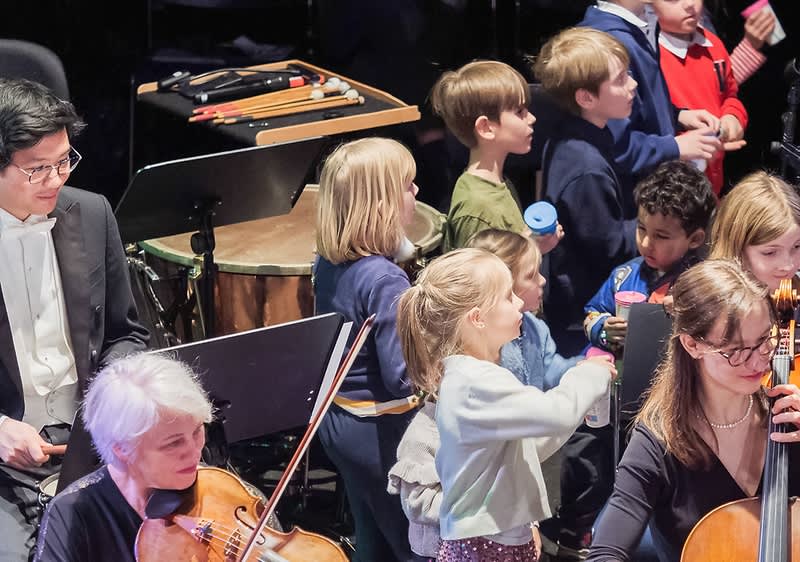 Eine Gruppe von Kindern in festlicher Kleidung steht auf einer Bühne, einige von ihnen tragen weiße Hemden oder Kleider, während im Vordergrund mehrere Hörner zu sehen sind, die von Musikern gespielt werden
