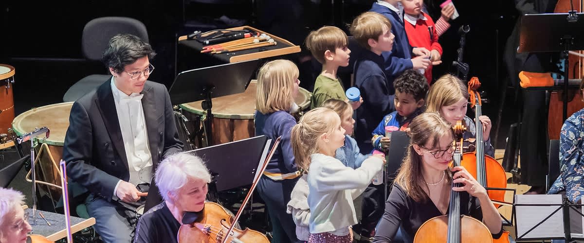Eine Gruppe von Kindern in festlicher Kleidung steht auf einer Bühne, einige von ihnen tragen weiße Hemden oder Kleider, während im Vordergrund mehrere Hörner zu sehen sind, die von Musikern gespielt werden