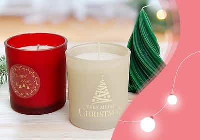 Zwei Kerzen auf einem Holztisch, links eine rote Kerze mit der Aufschrift „Home's Fest“, rechts eine weiße Kerze mit der Aufschrift „Very Merry Christmas“, im Hintergrund eine grüne, spiralförmige Kerze