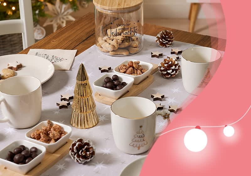 Ein festlich gedeckter Tisch mit weißen Tassen, kleinen Schalen mit Nüssen und Schokolade, einem goldenen Tannenbaum-Dekor, Tannenzapfen und einem Glasbehälter mit der Aufschrift „Wunder der Weihnachtszeit“