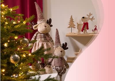 Zwei Stofftiere in Form von Elchen mit rosa Zipfelmützen und karierten Kleidern stehen neben einem geschmückten Weihnachtsbaum, im Hintergrund sind Holzfiguren von einem Hund mit rotem Schal und einem Tannenbaum auf einem Regal zu sehen