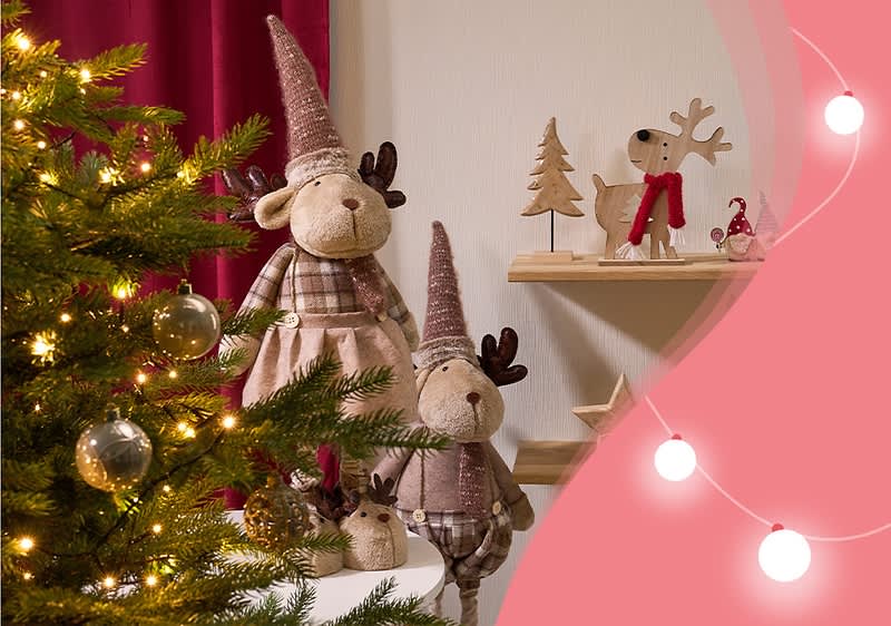 Zwei Stofftiere in Form von Elchen mit rosa Zipfelmützen und karierten Kleidern stehen neben einem geschmückten Weihnachtsbaum, im Hintergrund sind Holzfiguren von einem Hund mit rotem Schal und einem Tannenbaum auf einem Regal zu sehen