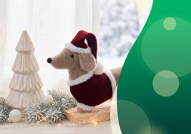 Plüschdackel mit rotem Weihnachtsmantel und Nikolausmütze steht auf einer Holzplatte neben einem weißen, gestuften Keramik-Weihnachtsbaum und frostigen Tannenzweigen mit Kugeln und Lichterkette. Bild mithilfe von KI erstellt