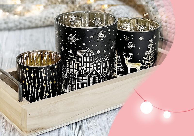 Drei schwarze Kerzenhalter mit goldenen winterlichen Motiven, darunter Schneeflocken, Häuser und ein Hirsch, stehen in einer Holzschale