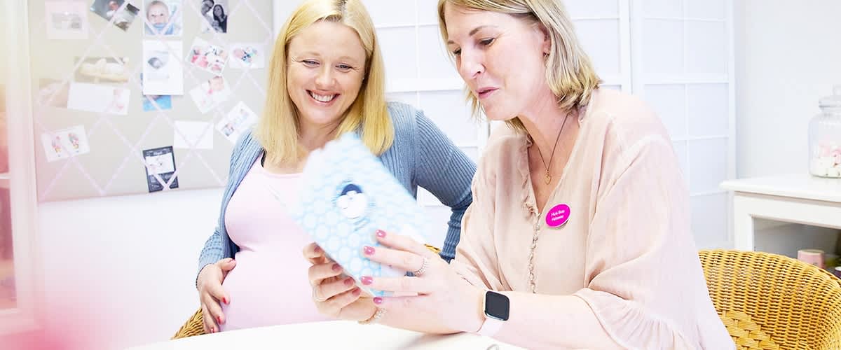 Zwei Frauen sitzen an einem Tisch, die eine Frau ist schwanger und trägt ein rosa Oberteil und eine blaue Strickjacke, während die andere Frau ein beiges Oberteil trägt und ein Namensschild mit einem pinken Aufkleber auf der Brust hat, beide halten ein Buch oder eine Broschüre mit einem Babyfoto darauf, im Hintergrund ist eine Pinnwand mit mehreren Babyfotos und Notizen zu sehen, auf dem Tisch stehen eine Tasse mit der Aufschrift Hallo Baby und eine rosa Schere