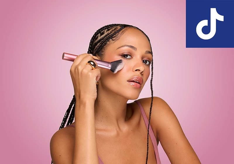 Frau mit geflochtenem Haar in pinkfarbenem Trägershirt hält ein roségoldenes Makeup-Produkt (zylindrisch) nahe ihres Gesichts vor ein einfarbiges rosa Hintergrundfeld, oben rechts blaues TikTok-Logo ohne Text