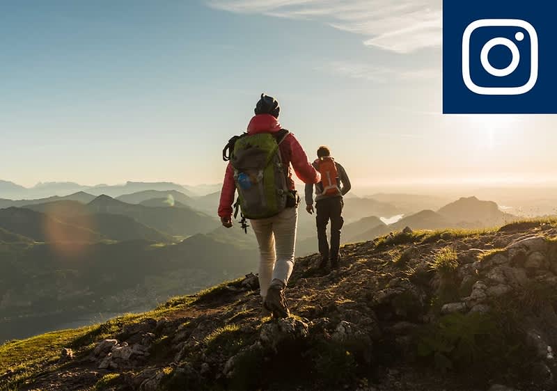 Zwei wandernde Personen mit Rucksäcken gehen auf einem felsigen Berggrat bei Sonnenaufgang, im Hintergrund Berge und Tallandschaft, oben rechts Instagram-Logo