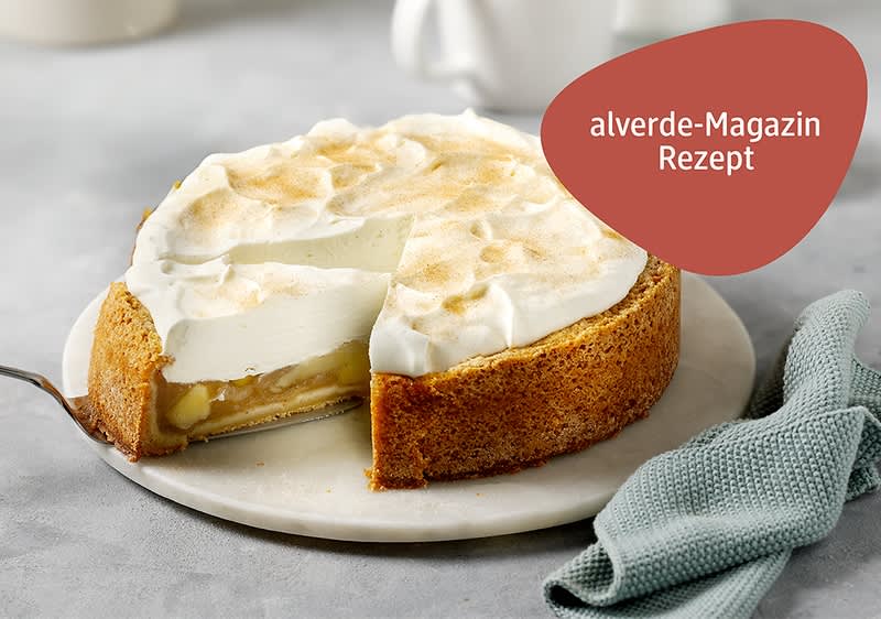 Eine Apfel-Schmand-Torte mit einer goldbraunen Kruste auf einem weißen Teller, daneben liegt ein hellgrünes, strukturiertes Tuch, im Hintergrund sind verschwommen eine weiße Teekanne und Tassen zu sehen.