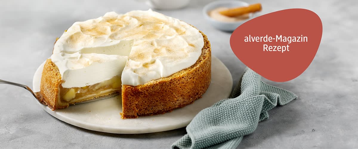 Eine Apfel-Schmand-Torte mit einer goldbraunen Kruste auf einem weißen Teller, daneben liegt ein hellgrünes, strukturiertes Tuch, im Hintergrund sind verschwommen eine weiße Teekanne und Tassen zu sehen.