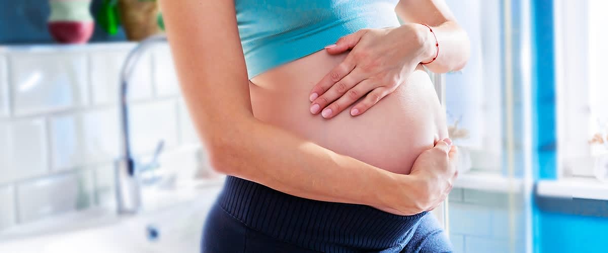 Eine schwangere Frau in einem blauen Top hält ihre Hände sanft auf ihren Bauch