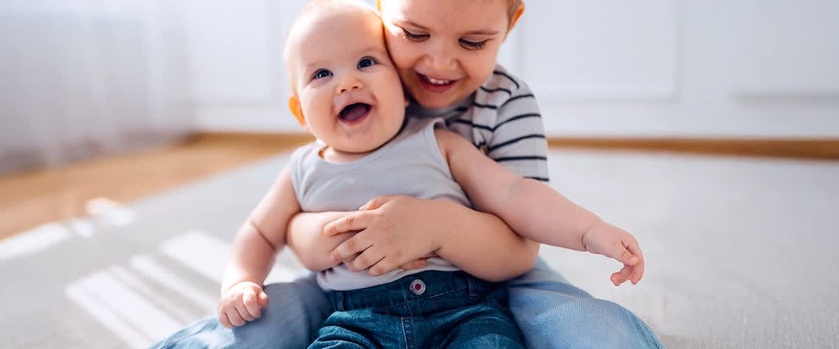 Ein Kleinkind in einem grauen Tanktop und blauer Hose sitzt auf dem Schoß eines älteren Kindes in einem gestreiften Shirt, beide haben verschwommene Gesichter