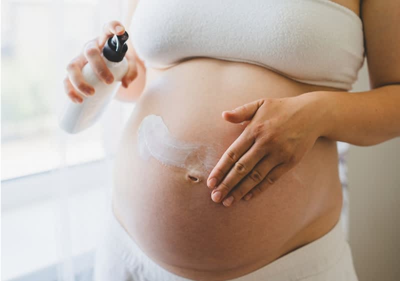 Schwangere Frau in weißem Top cremt ihren nackten runden Bauch mit Lotion aus einer Pumpflasche
