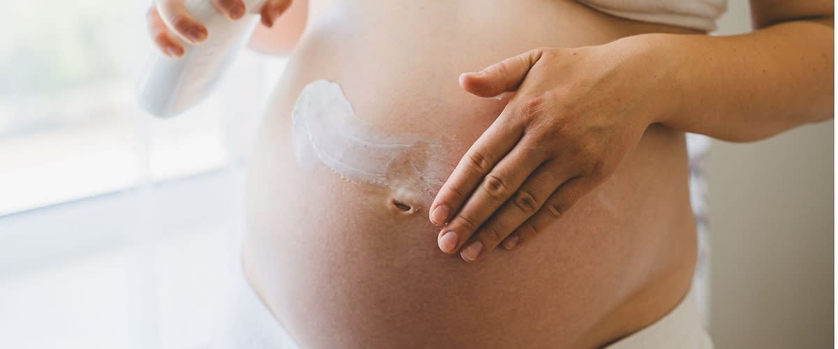 Schwangere Frau in weißem Top cremt ihren nackten runden Bauch mit Lotion aus einer Pumpflasche