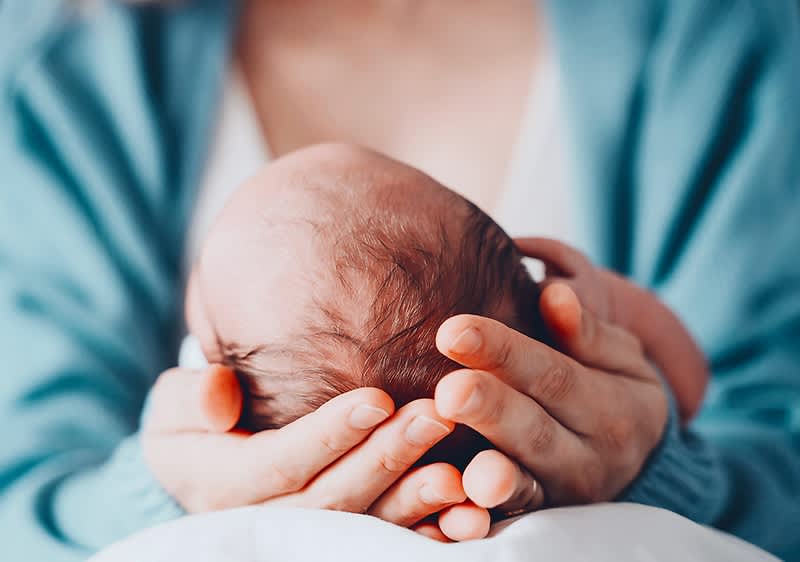 Eine Person in einem hellblauen Pullover hält sanft ein Neugeborenes in ihren Händen, wobei das Baby mit dem Kopf nach unten liegt und die Hände der Person das Baby sicher umschließen, während der Hintergrund unscharf ist und die Szene eine ruhige und liebevolle Atmosphäre vermittelt.