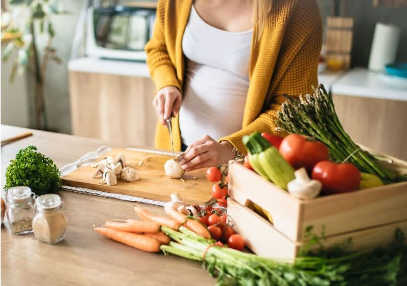 Frau in gelbem Cardigan schneidet auf einem Holzbrett eine Champignon, vor ihr liegen Karotten, Kirschtomaten, Sellerie, Spargel und eine Holzkiste mit Tomaten und anderem Gemüse