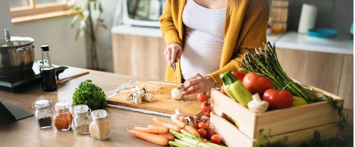 Frau in gelbem Cardigan schneidet auf einem Holzbrett eine Champignon, vor ihr liegen Karotten, Kirschtomaten, Sellerie, Spargel und eine Holzkiste mit Tomaten und anderem Gemüse
