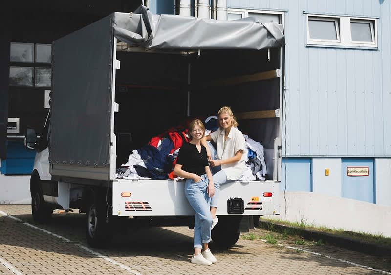 Zwei junge Frauen stehen lächelnd vor einem offenen Lieferwagen, der mit einer großen Menge an Kleidung beladen ist, die in verschiedenen Farben und Stoffen sichtbar ist, im Hintergrund ist ein blaues Gebäude mit Fenstern und einer Tür zu sehen, die Frauen tragen lässige Kleidung, eine in einem schwarzen Oberteil und Jeans, die andere in einem weißen Oberteil und heller Hose, der Boden ist mit Pflastersteinen bedeckt