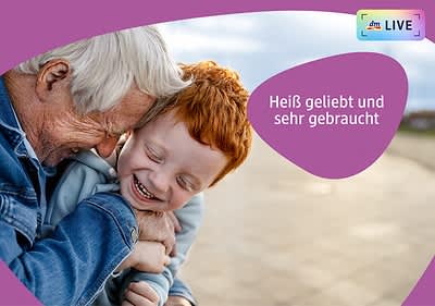 Ein älterer Mann mit grauem Haar und ein Kind mit roten Haaren umarmen sich innig, beide tragen blaue Kleidung, der Hintergrund zeigt einen unscharfen Außenbereich, oben rechts steht in weißer Schrift auf einem lila Hintergrund Heiß geliebt und sehr gebraucht.