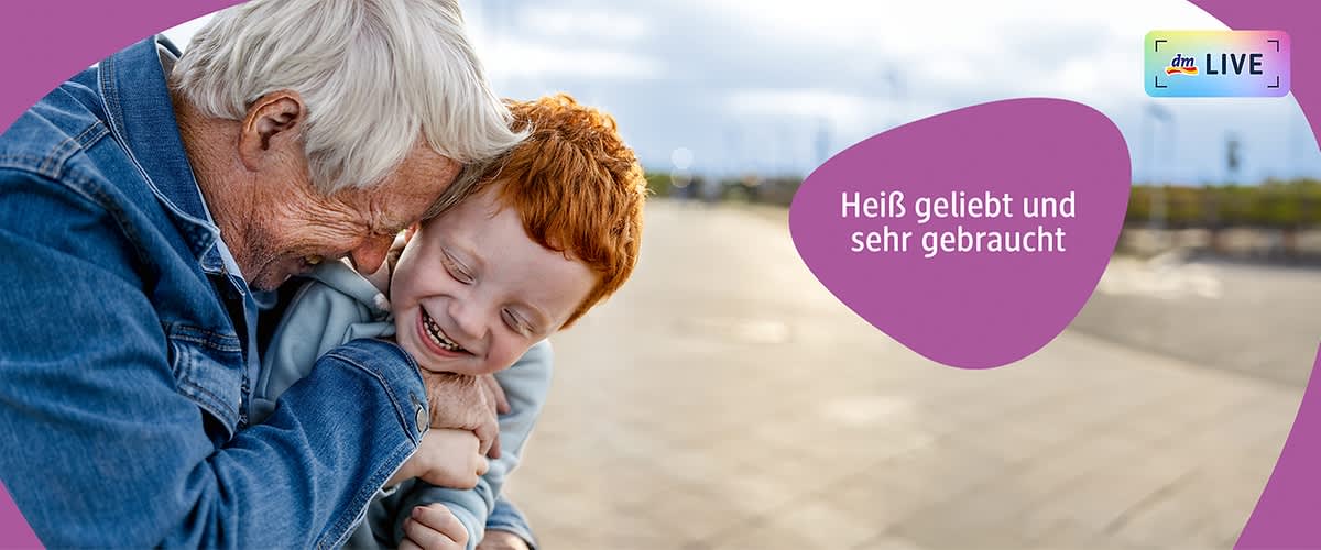 Ein älterer Mann mit grauem Haar und ein Kind mit roten Haaren umarmen sich innig, beide tragen blaue Kleidung, der Hintergrund zeigt einen unscharfen Außenbereich, oben rechts steht in weißer Schrift auf einem lila Hintergrund Heiß geliebt und sehr gebraucht.
