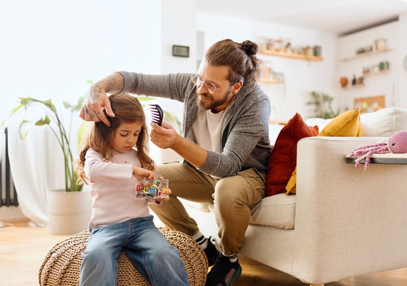 Ein Mann kämmt die Haare eines kleinen Mädchens, das auf einem runden Hocker sitzt und ein Spielzeug hält, im Hintergrund ein helles Wohnzimmer mit Pflanzen und einem Sofa mit bunten Kissen