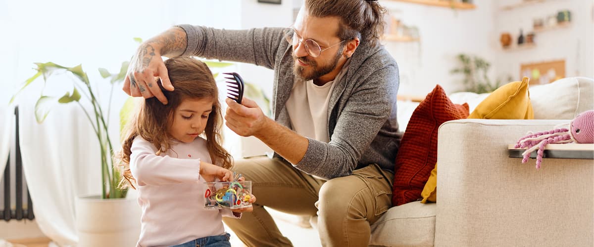 Ein Mann kämmt die Haare eines kleinen Mädchens, das auf einem runden Hocker sitzt und ein Spielzeug hält, im Hintergrund ein helles Wohnzimmer mit Pflanzen und einem Sofa mit bunten Kissen