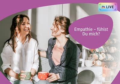 Zwei Personen sitzen nebeneinander auf einem Sofa und halten jeweils eine Tasse, die linke Person trägt ein weißes Hemd und hält eine grüne Tasse, die rechte Person trägt ein dunkelblaues Hemd und hält eine rote Tasse mit weißen Punkten, im Hintergrund sind mehrere Tassen und eine Kaffeemaschine zu sehen, oben rechts steht der Text „Empathie – fühlst Du mich?“