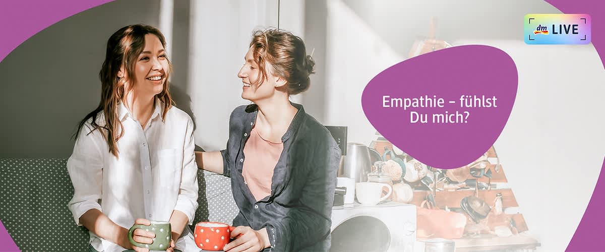 Zwei Personen sitzen nebeneinander auf einem Sofa und halten jeweils eine Tasse, die linke Person trägt ein weißes Hemd und hält eine grüne Tasse, die rechte Person trägt ein dunkelblaues Hemd und hält eine rote Tasse mit weißen Punkten, im Hintergrund sind mehrere Tassen und eine Kaffeemaschine zu sehen, oben rechts steht der Text „Empathie – fühlst Du mich?“