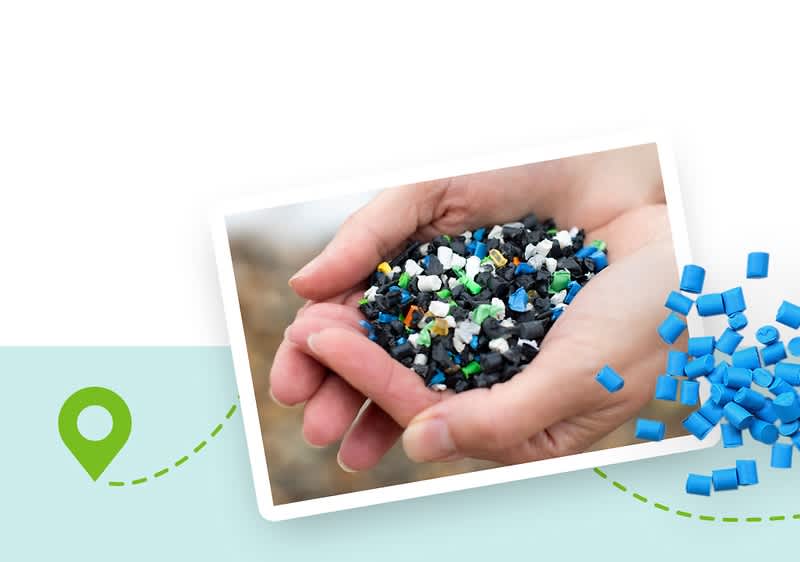 Zwei Hände halten eine Mischung aus kleinen Plastikstücken in verschiedenen Farben, darunter Schwarz, Weiß, Blau und Grün, daneben liegen blaue Plastikpellets auf einer hellblauen Fläche mit einem grünen Standort-Symbol und einer gestrichelten Linie