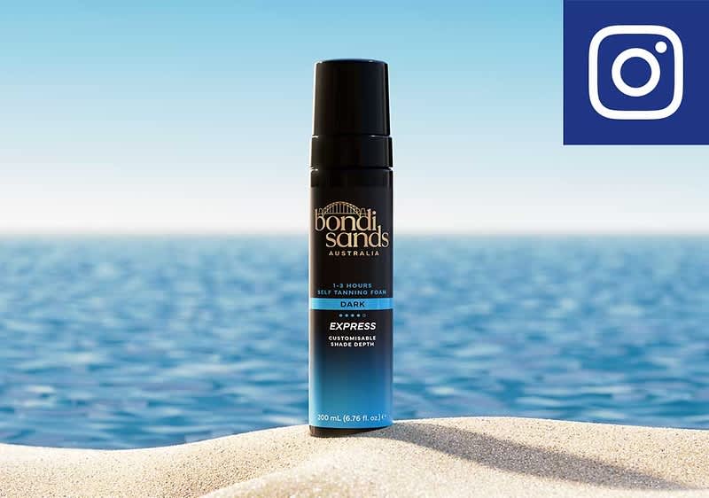 Eine schwarze und blau gestaltete Sprühflasche von „Bondi Sands“ mit der Aufschrift „Express Foam Shave Gotta“ steht auf hellem Sand vor unscharfem Meeres- und Himmelshintergrund und oben rechts ein blaues Quadrat mit weißem Instagram-Logo